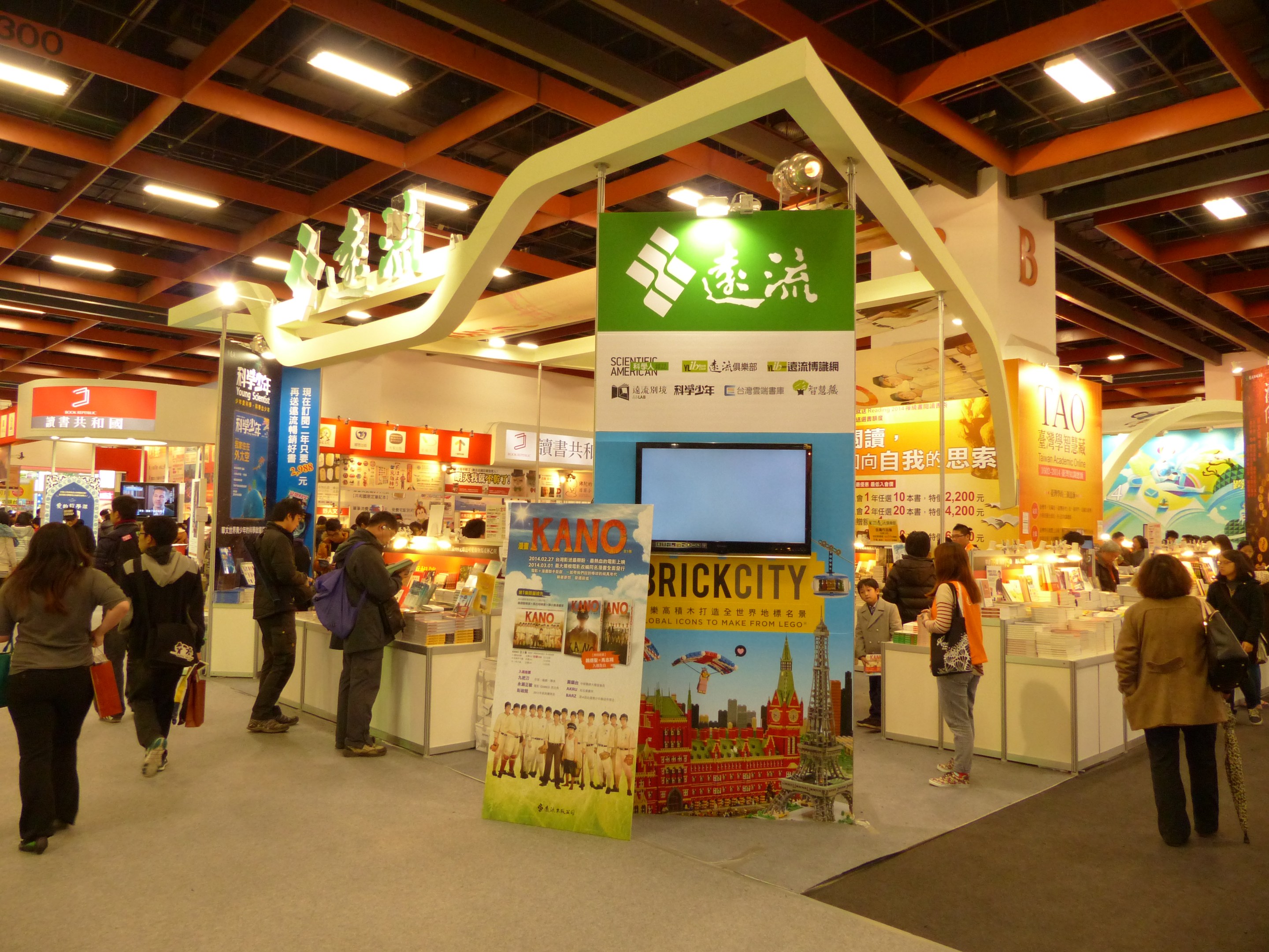 Eine Gruppe von Menschen vor einem Messestand mit Bannern, Fernseher, Tischen mit Gegenständen, Schildern und Deckenleuchten