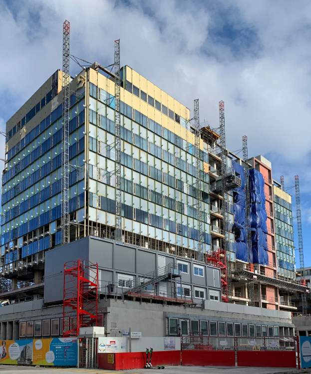 Großes Gebäude im Bau in einer städtischen Umgebung, das Glasfenster, Metallrahmen, eine Treppe, einen Zaun, ein Schild, Pfosten, einen Straßenpfosten, umgebende Bäume und einen bewölkten Himmel zeigt.