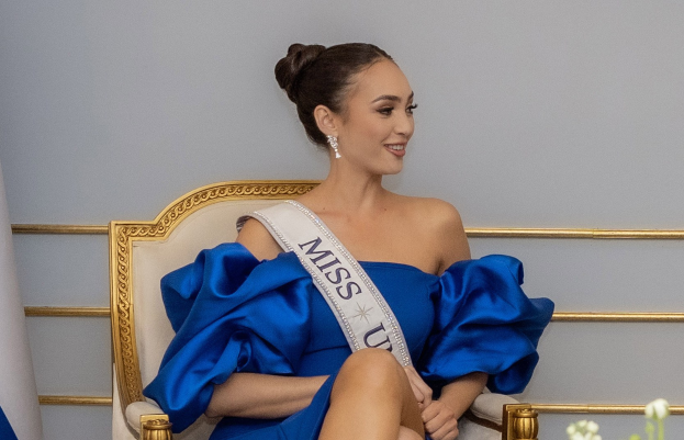 Frau in einem blauen Kleid mit einem 'Miss Universe 2019'-Band, die auf einem Stuhl sitzend von Blumen umgeben ist und lächelt.