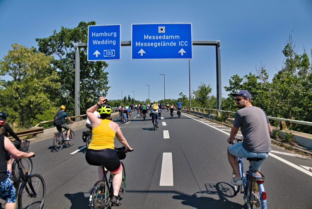 Eine Gruppe von Menschen in Helmen fährt auf Fahrrädern eine Straße mit einer Begrenzung auf einer Seite und Bäumen auf der anderen, mit Laternen und einem klaren blauen Himmel im Hintergrund und einer Tafel oben, die eine Fahrradtour in Hamburg anzeigt.