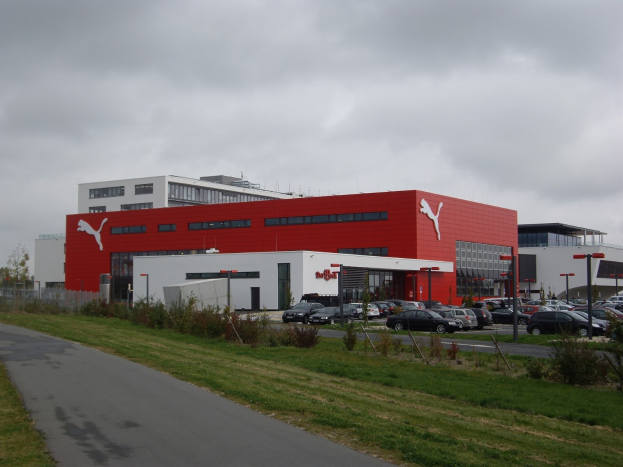 Übersichtsansicht der Puma-Fabrik in Deutschland mit Gebäuden, Fahrzeugen, Straßeninfrastruktur, Vegetation und einem bewölktem Himmel.