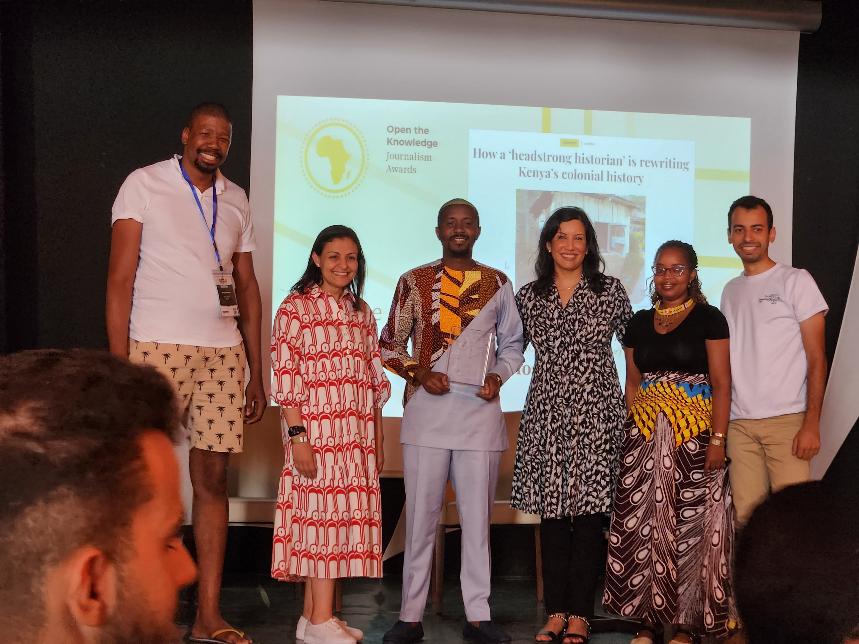 Gruppe von Menschen, die auf einer Bühne stehen, lächeln und für ein Foto posieren, mit sitzendem Publikum darunter und einem Bildschirm im Hintergrund, der "Open the Knowledge Journalism Awards 2019" anzeigt.