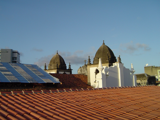 Stadtansicht mit mehreren Gebäuden im Vordergrund unter einem blauen Himmel, mit Solarpanelen auf dem Dach eines Gebäudes.