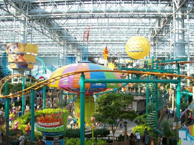 Indoor-Wasserpark mit einer Achterbahn, umgeben von Menschen, Bäumen, Pflanzen und deckenmontierten Stangen mit Lichtern.