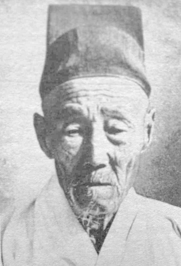 Schwarzes und weißes Foto eines alten Mannes in einer Mütze und einem Kimono, der vor einer Wand steht.