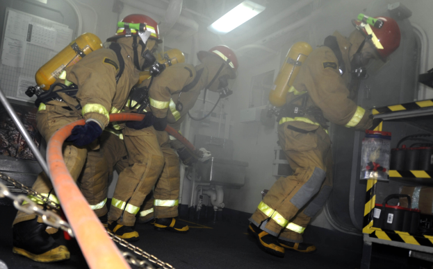 Feuerwehrleute in Schutzausrüstung arbeiten an einem Löschfahrzeug mit Ausrüstungsgestellen und Hintergrundinfrastruktur.
