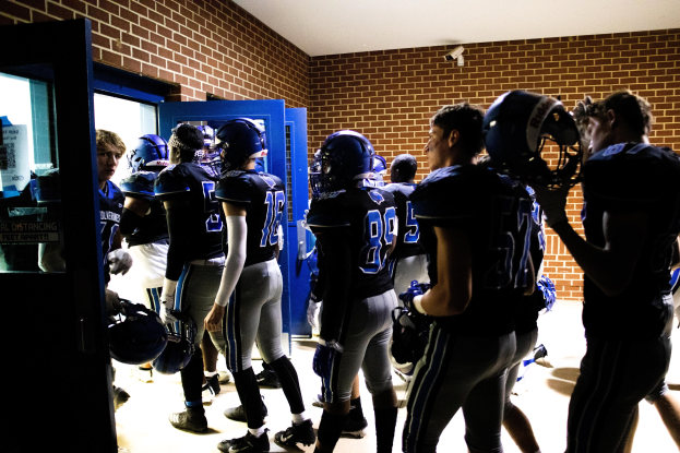 Gruppe von Fußballspielern in Uniformen und Helmen, die in einer Umkleidekabine mit sichtbaren Wänden und einer Tür auf der linken Seite stehen.