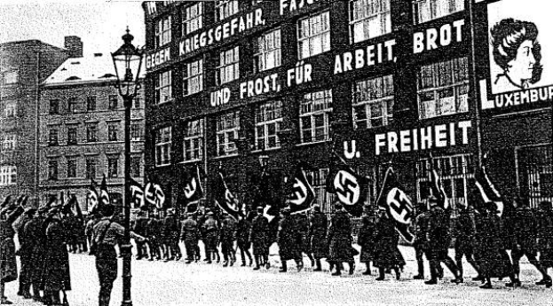 Schwarzes und wei├čes Bild einer Gruppe, die mit Fahnen und Spruchb├Ąndern die Stra├če entlangmarschiert, mit einem Lichtmast links und einem Gebäude mit einem Nazi-Parteischild im Hintergrund.