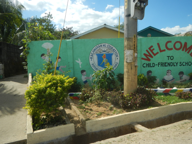 Eine grüne Wand mit einem Schild, auf dem "Willkommen in der kinderfreundlichen Schule" steht, umgeben von Häusern, Bäumen, Pflanzen und einem Mast mit einer Lampe.