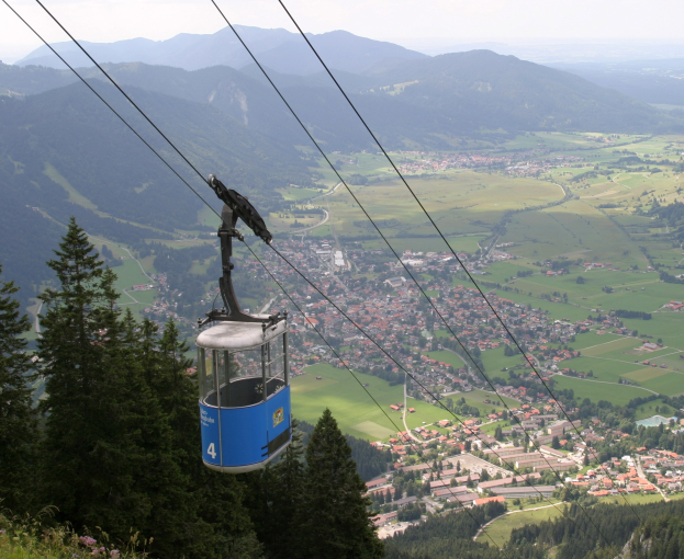 Eine Seilbahn fährt einen bewaldeten Berg hinauf, mit einer kleinen Stadt unten und sanften Hügeln unter einem klaren blauen Himmel.