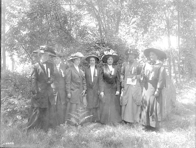 Eine Gruppe von Frauen in Hüten, die zusammen in einem bewaldeten Gebiet stehen, gekleidet als Suffragetten, auf einem Schwarz-Weiß-Bild.