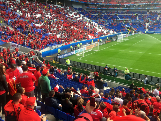 Eine große Menschenmenge in einem Stadion schaut einem Fussballspiel zu, mit Menschen auf Sitzplätzen und Stehplätzen, Fahnen, einem Torpfosten und Deckenleuchten.