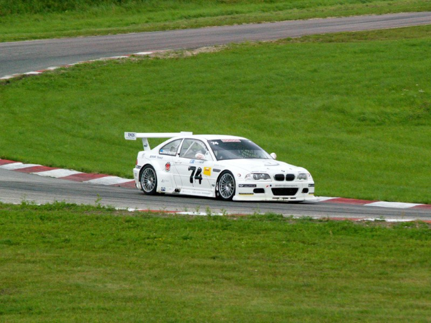 Weißer BMW M3 GT3 Rennwagen auf einer Rennstrecke mit grünem Gras und Pflanzen im Hintergrund.