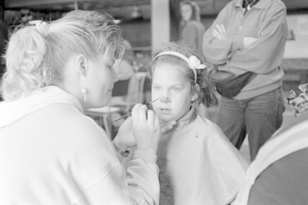 Ein junges Mädchen lässt sich von einer Frau im Salon schminken, während zwei andere Personen im Hintergrund stehen, alles in Schwarz-Weiß.