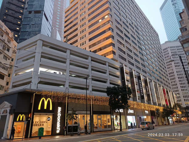 Ein McDonald's-Restaurant auf einer belebten Stadtstraße, umgeben von hohen Gebäuden, Straßenlaternen, Straßenlaternen, fahrenden Fahrzeugen, Fußgängern, Bäumen und einem klaren blauen Himmel.