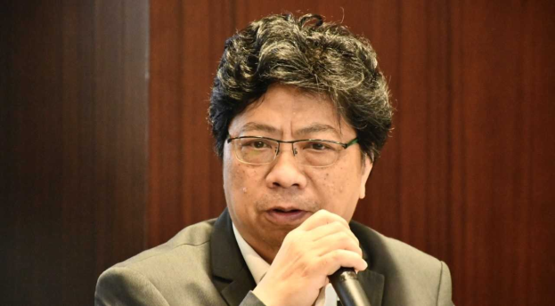 Ein Mann im Anzug mit einem Mikrofon, der lebhaft vor einer hölzernen Wand spricht.