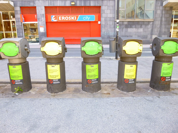 Reihe von Parkautomaten entlang einer Straße mit einem Gebäude mit Glasfenstern und einer Rolllade mit Text sowie einem nahen Mülleimer.