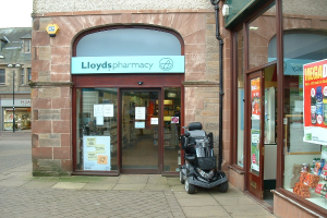 Eine Apothekenfiliale mit einem vor ihr geparkten Fahrzeug und einem Gebäude in der linken Ecke.