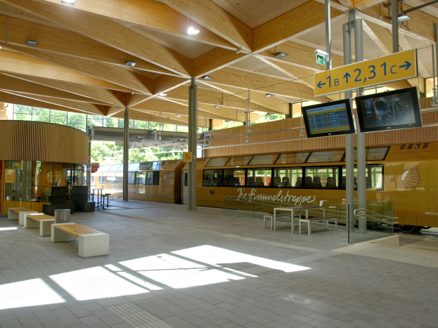Ein belebter Bahnhof mit einem Zug auf den Schienen, umgeben von Bänken, Tischen, Mästen, Schildern, Lämpchen und Bäumen, wo Menschen auf den Zug warten.
