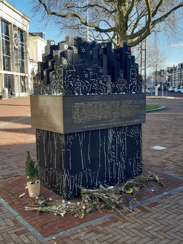 Denkmal in einem Stadtplatz für Holocaust-Opfer, mit einem Baum davor, einem Topf mit einer Pflanze darin, verstreuten trockenen Blättern und Gebäuden, Fahrzeugen und Bäumen im Hintergrund.