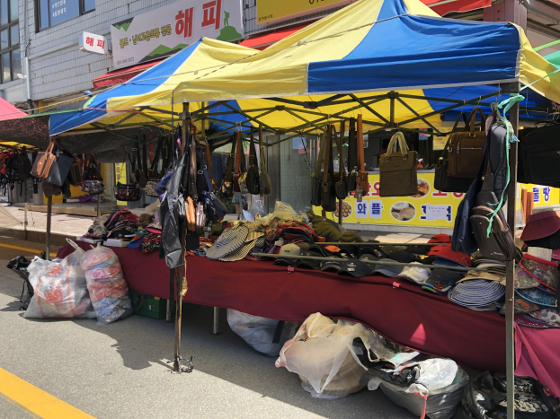 Ein Markt mit Ständen, die Taschen, Hüte und andere Gegenstände präsentieren, vor einem Hintergrund von Gebäuden mit Fenstern und Texttafeln.