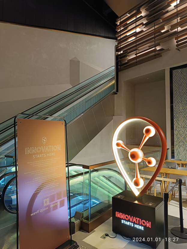 Großes Schild in einem Gebäude mit einer Rolltreppe im Hintergrund, Tische und Stühle auf der rechten Seite und eine Tafel mit der Aufschrift "Innovation Starts Here" auf der linken Seite, was die Teilnahme an der HSTP20 Expo anzeigt.