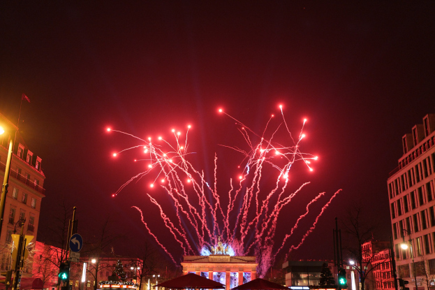 Eine belebte Stadtstraße bei Nacht am Silvesterabend in Berlin, mit Gebäuden, Bäumen, Laternenmasten, Verkehrszeichen, Zeltplanen, Menschen und einem farbenfrohen Feuerwerk, das den Himmel erhellt.