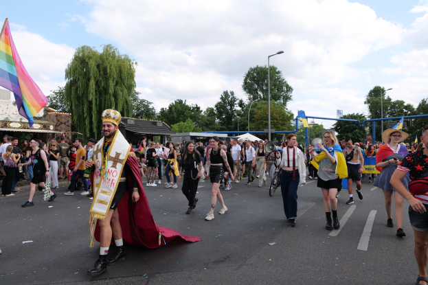 Eine Gruppe von Menschen marschiert bei der Pride Parade 2018 mit einer Regenbogenflagge und Musikinstrumenten, während im Hintergrund Laternenpfähle, Bäume und Hütten zu sehen sind.