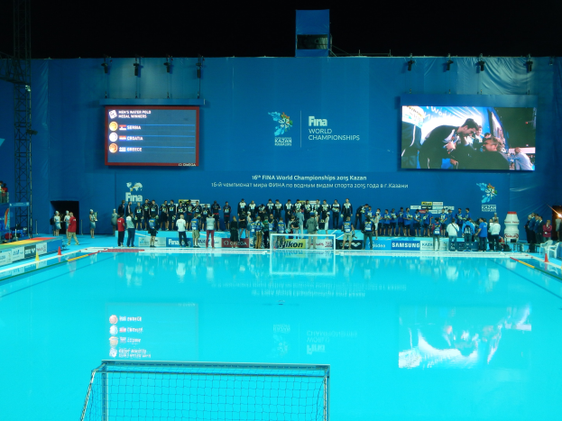 Gruppe von Menschen um ein Hallenbad mit Netz am Boden herumstehend, mit einer Wand mit "FINA World Championships 2015 Kazan"-Text und -Logos, Trägern und Lichtern im Hintergrund.