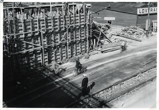 Schwarz-weiß-Foto einer Baustelle mit einem teilweise errichteten Gebäude, Menschen auf Fahrrädern und zu Fuß auf der Straße, Bahnschienen, Pfosten, Bretter und Text unten.