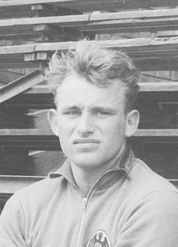 Schwarzes und weißes Foto eines Mannes in einer Jacke, der vor gestapelten Holzplanken steht, der angeblich ein legendärer deutscher Fußballspieler ist, der im Alter von 89 Jahren verstorben ist.