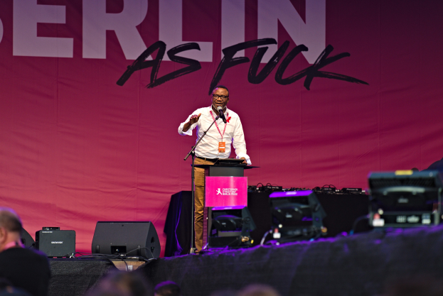 Ein Mann in einem weißen Hemd und braunen Hosen steht auf einer Bühne und hält ein Mikrofon, während er eine Menge anspricht, mit einem pinken Banner mit der Aufschrift "Berlin as Fuck" im Hintergrund und sichtbarer Bühnenausrüstung.