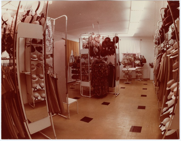 Altes Foto eines Kleiderladen-Interieurs mit Kleidung an Bügeln, Hüten auf Ständern, einem Spiegel, einem Stuhl, einem Tisch mit Gegenständen, Vorhängen und Deckenlampen.