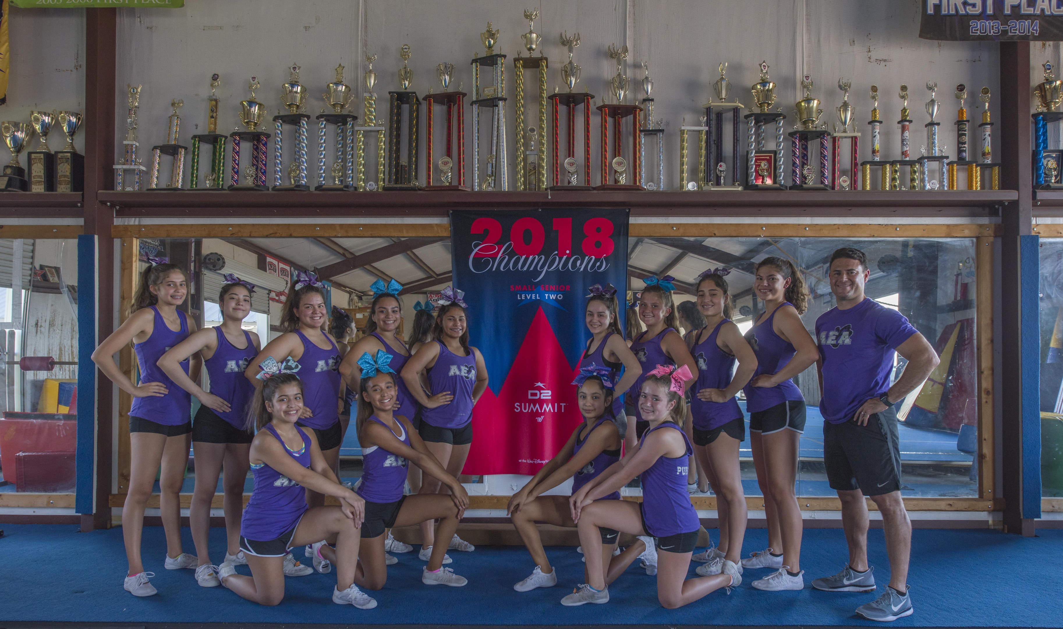Gruppe von Cheerleadern in lila T-Shirts und schwarzen Shorts, die vor einem Vitrinenschrank mit einer '2018 Summit Cheerleading Champions'-Aufschrift posieren.