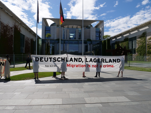Eine Gruppe von Menschen hält ein Transparent mit der Aufschrift 'Deutschland, Lagerland Migration ist kein Verbrechen' vor einem umzäunten Gebäude mit Bäumen und Flaggen.