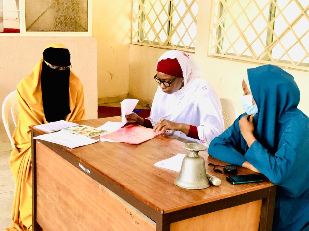 Drei Frauen in einem raumähnlichen Setting sitzen an einem Tisch mit Papieren, Büchern, einer Glocke und einem Mobiltelefon und diskutieren über Geschlechtergerechtigkeit in Nigeria.