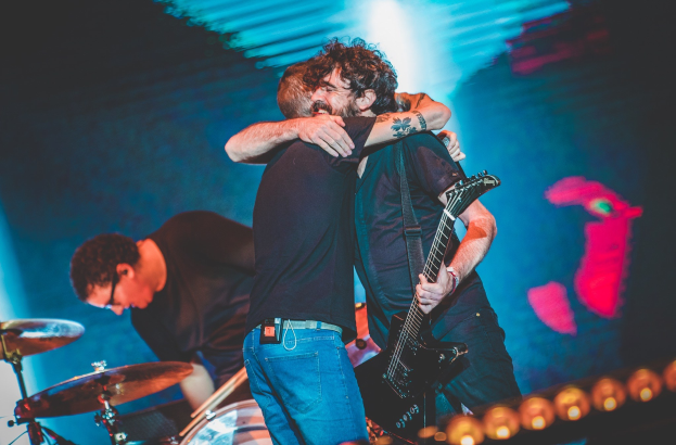 Zwei Männer umarmen sich auf der Bühne eines Konzerts, wobei einer eine Gitarre spielt und ein anderer am Schlagzeug sitzt, beleuchtet von Scheinwerfern.