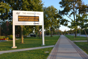 Ein Schild am Boden mit der Aufschrift "Die Versammlungshalle - Treffen Sie Ihre Landtagswahl-Kandidaten", umgeben von Gras, einem Weg, Bäumen, blühenden Pflanzen, Straßenlaternen, Fahrzeugen, einem Gebäude mit Fenstern und einem bewölkten Himmel.
