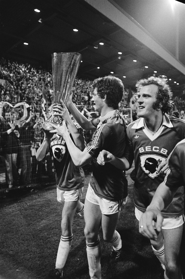 Schwarzes Bild von Fußballspielern, die mit einem Pokal auf einem Feld feiern, während Zuschauer hinter einem Zaun zu sehen sind, unter Stadionbeleuchtung.