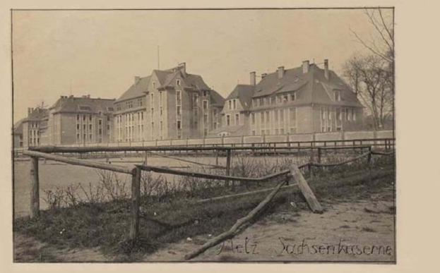 Ein altes Schwarz-Weiß-Foto eines großen Gebäudes mit Fenstern, umgeben von einem Zaun, Gras, Pflanzen, Bäumen und einem Himmel im Hintergrund, mit dem Text "Sachsenhausen, Deutschland" unten.