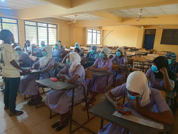 Eine Klasse in Nigeria mit Schülern, die Gesichtsmasken tragen und auf Bänken sitzen oder stehen, mit Büchern und Gegenständen auf Tischen, Fenstern, einer Tür und Deckenleuchten und -lüftern im Hintergrund.