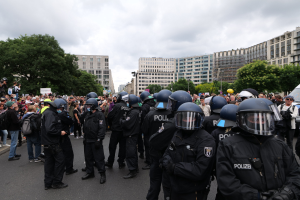 Eine große Gruppe von Polizisten steht vor einer Menschenmenge auf einer Straße, umgeben von Bäumen und Gebäuden mit Fenstern, mit einem bewölkten Himmel im Hintergrund; einige in der Menge halten Kameras während einer Demonstration in Berlin, Deutschland.