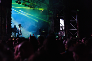 Eine Menge schaut Performern auf einer hell erleuchteten Bühne bei einem Konzert zu, mit Musikinstrumenten, Mikrofonen und einem Banner.