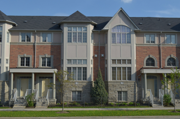 Eine Reihe von Stadth├Ąusern in einer Wohngegend mit sichtbaren Fenstern, Türen, Pfeilern, Gel├Ąndern, Stufen, Pflanzen, B├Ąumen, Gras, einer Stra├če im Vordergrund und Himmel im Hintergrund.