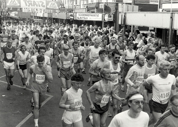 Eine große Gruppe von Menschen, die bei einem Marathon auf einer Straße laufen, umgeben von Gebäuden, Polen, Plakaten und anderen Gegenständen, dargestellt in Schwarz-Weiß.