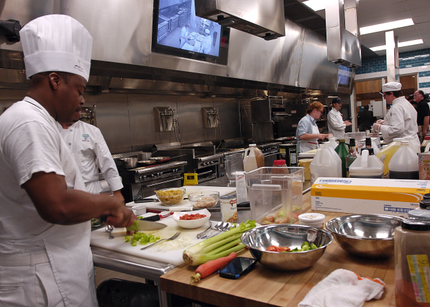 Eine Gruppe von Köchen in einer Küche, die Essen auf einem Schneidebrett zubereiten, wobei ein Koch in der vorderen Reihe in einer weißen Schürze und einer Mütze steht und ein Messer hält und von Utensilien, Schüsseln, Gemüse, einem Mobiltelefon, Flaschen und anderen Gegenständen umgeben ist, mit einem Fernseher und Deckenlampen im Hintergrund.