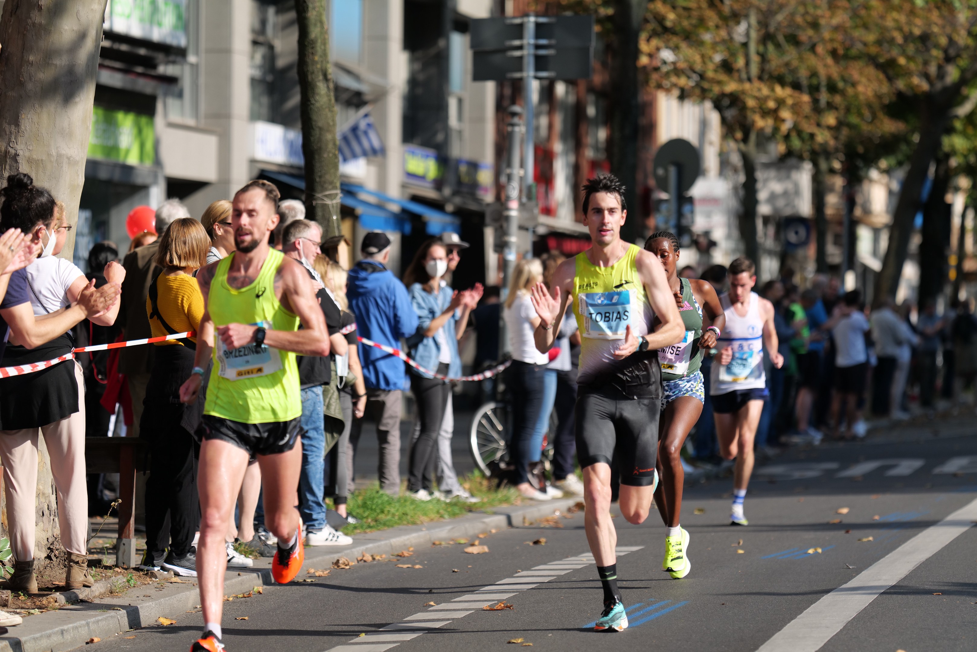 Gruppe von Läufern bei einem Marathon auf einer Stadtstraße, mit Zuschauern auf dem linken Gehweg und Gebäuden, Bäumen und einem klaren blauen Himmel im Hintergrund.