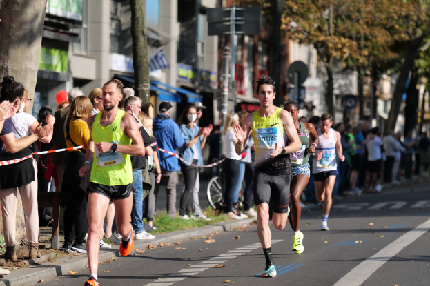 Gruppe von Läufern bei einem Marathon auf einer Stadtstraße, mit Zuschauern auf dem linken Gehweg und Gebäuden, Bäumen und einem klaren blauen Himmel im Hintergrund.