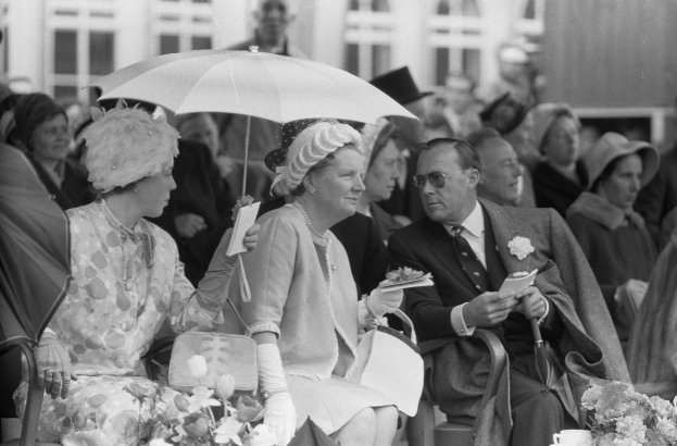 Schwarzes und weißes Foto von Queen Elizabeth II und Prinz Philip, die mit Schirmen sitzen, umgeben von einer Gruppe von Menschen mit Hüten und Büchern, mit einem Gebäude und Blumenarrangements im Hintergrund.