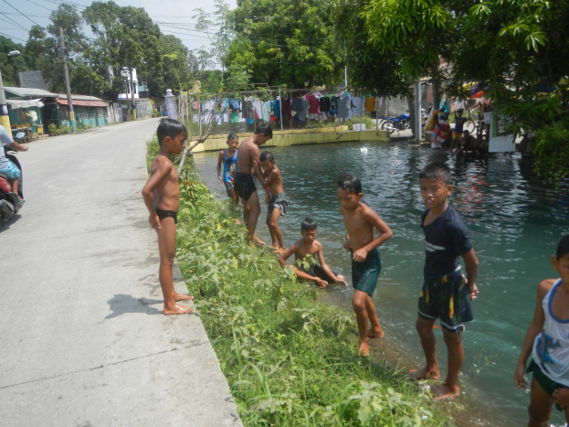 Kinder beim Spielen im flachen Wasser am Straßenrand, einige sitzen auf dem Gras, eine Person auf einem Motorrad links und Häuser, Bäume und einen klaren blauen Himmel im Hintergrund.